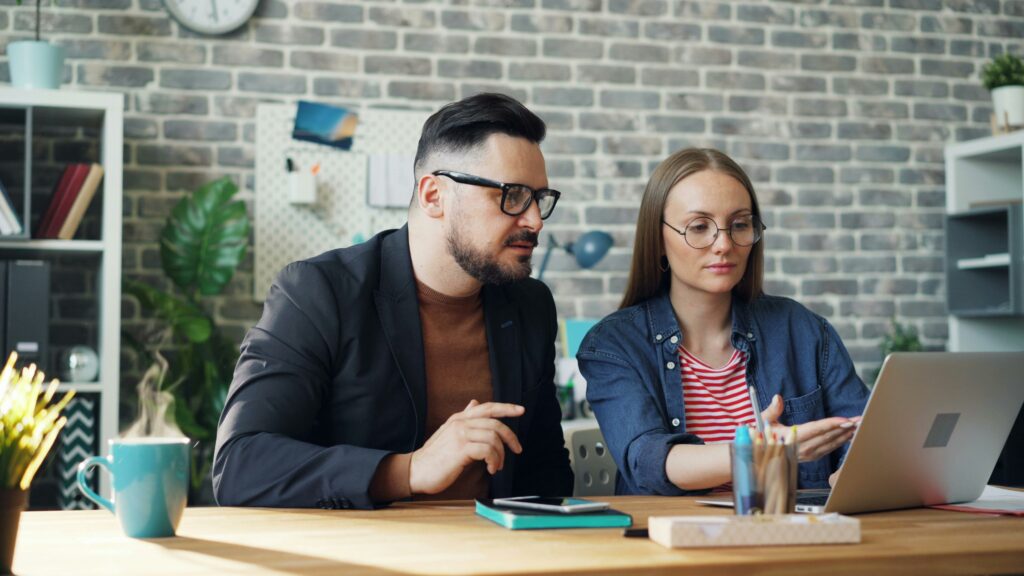 A man and a woman sit at a table in an office, looking at a laptop screen in front of the woman.